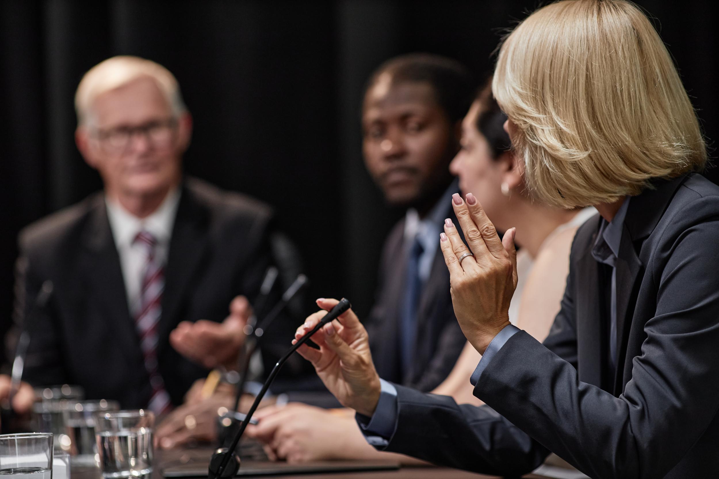 A woman speaking into a desk microphone at a long table of people